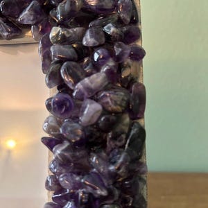 May include: A rectangular picture frame featuring a border densely covered in small, polished, deep purple amethyst stone chips. The stones vary in size and shape, creating a textured, multi-faceted surface that reflects light across the frame edge.