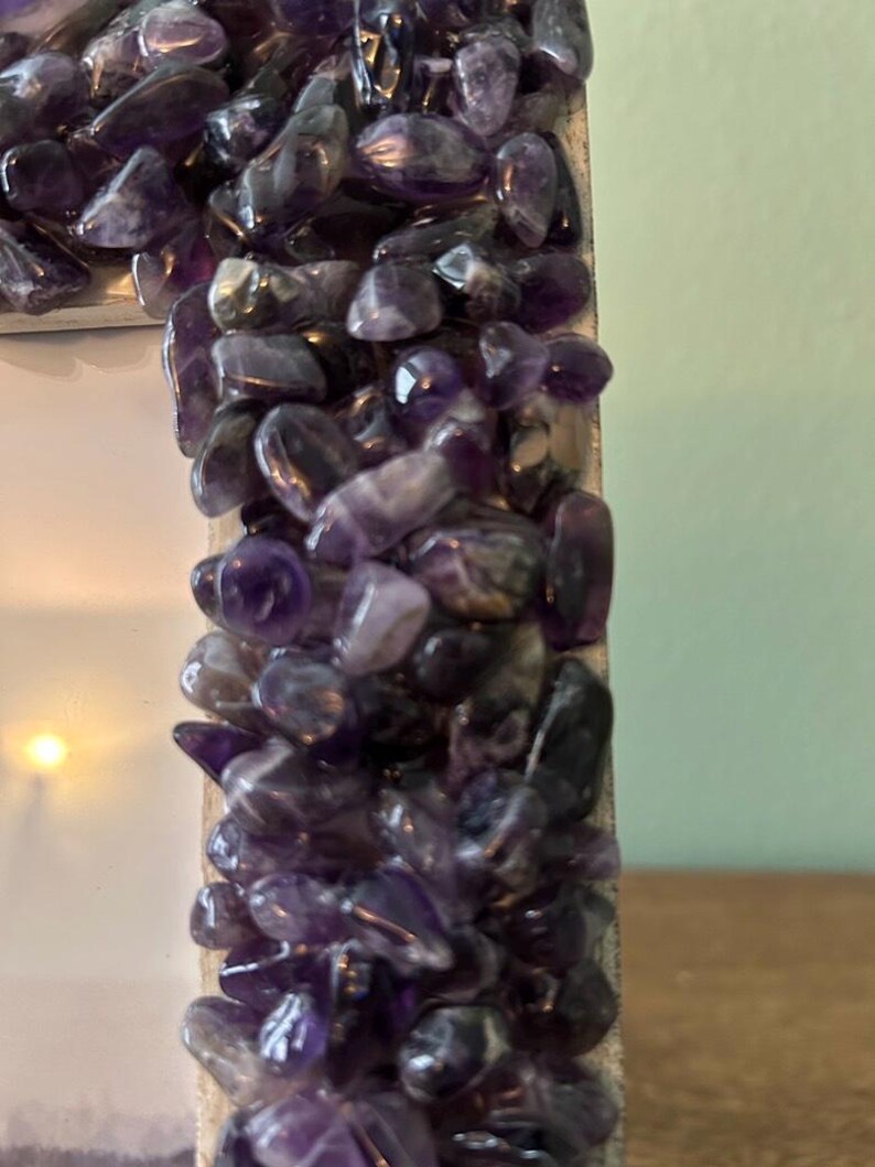 May include: A rectangular picture frame featuring a border densely covered in small, polished, deep purple amethyst stone chips. The stones vary in size and shape, creating a textured, multi-faceted surface that reflects light across the frame edge.