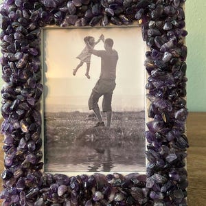 May include: A rectangular picture frame featuring a border densely covered in small, polished purple amethyst stones. The frame holds a black and white photograph depicting an adult lifting a child into the air in an outdoor setting near water.