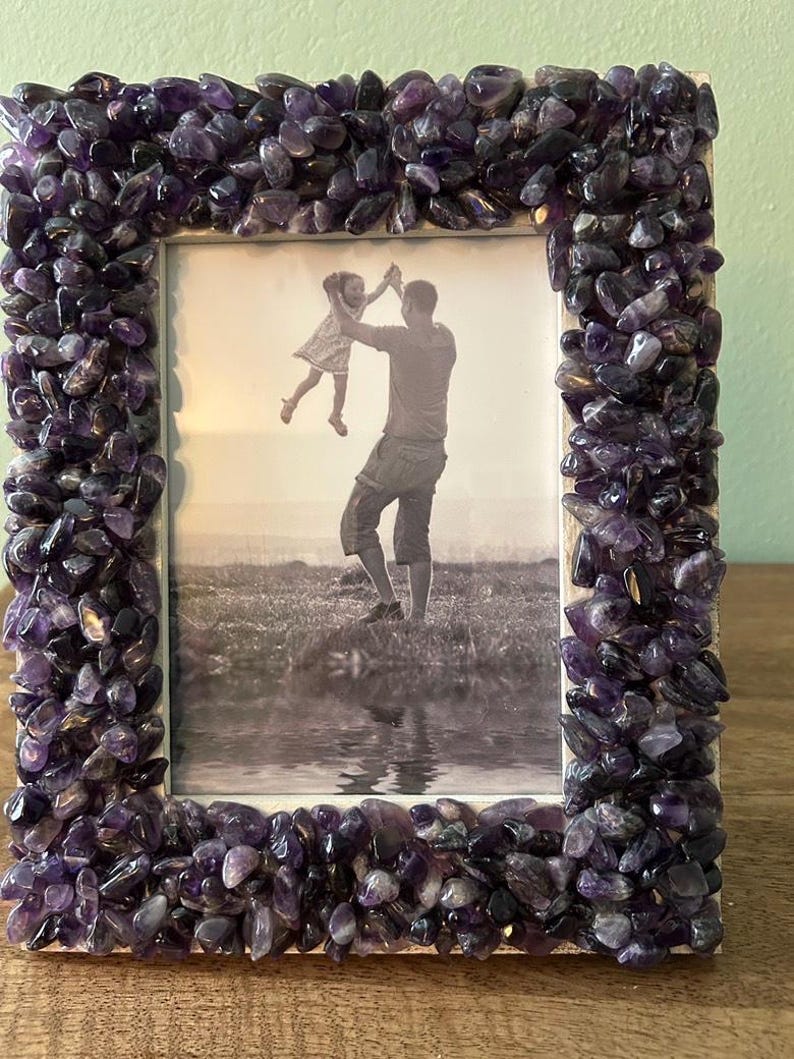 May include: A rectangular picture frame featuring a border densely covered in small, polished purple amethyst stones. The frame holds a black and white photograph depicting an adult lifting a child into the air in an outdoor setting near water.