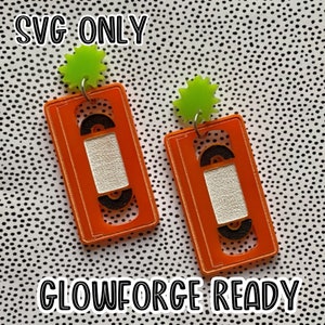 Puede incluir: Un par de pendientes de cassette naranja y negro con abalorios en forma de estrella verde. Los pendientes están listos para ser cortados en una Glowforge.