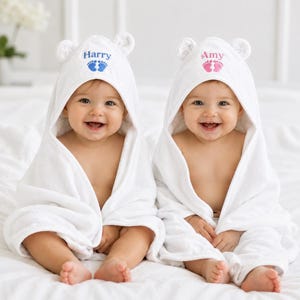 May include: Two babies wearing white hooded towels with bear ears. One towel has the name "Harry" and blue footprints, the other has "Amy" and pink footprints. Both babies are smiling.