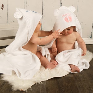 May include: Two white hooded baby towels with bear ears. One towel has the name "Amy" and pink footprints embroidered on the hood. The other towel has blue embroidery. The babies are sitting on a white faux fur rug.