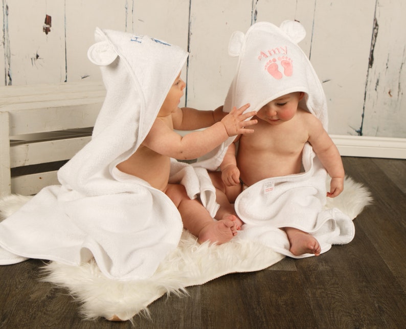 May include: Two white hooded baby towels with bear ears. One towel has the name "Amy" and pink footprints embroidered on the hood. The other towel has blue embroidery. The babies are sitting on a white faux fur rug.