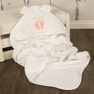 May include: A white hooded baby towel with bear ears. The hood is embroidered with the name "Amy" in pink, along with two pink baby footprints. The towel is folded and draped over a white wooden crate.