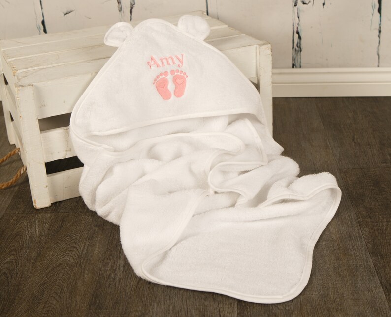 May include: A white hooded baby towel with bear ears. The hood is embroidered with the name "Amy" in pink, along with two pink baby footprints. The towel is folded and draped over a white wooden crate.
