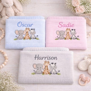 May include: Three folded baby blankets in light blue, light pink, and light grey. Each blanket features an embroidered design of a cartoon elephant, giraffe, and zebra sitting in grass. The names Oscar, Sadie, and Harrison are embroidered above.