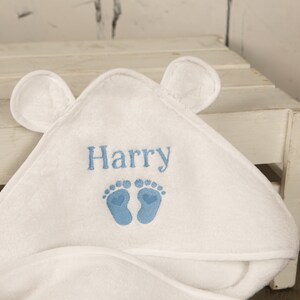 May include: White hooded baby towel with embroidered name "Harry" in blue, along with blue footprints. The towel has two small, rounded ears on the hood.