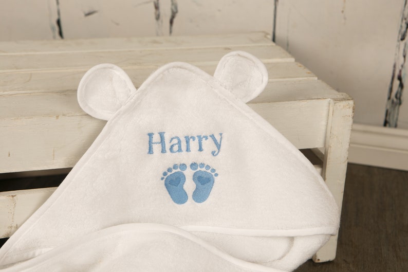 May include: White hooded baby towel with embroidered name "Harry" in blue, along with blue footprints. The towel has two small, rounded ears on the hood.