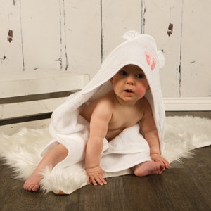 May include: A baby wrapped in a white hooded towel with pink footprints on the hood. The baby is sitting on a white faux fur rug. The background includes a white wooden crate and a white wooden wall.