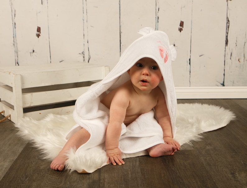 May include: A baby wrapped in a white hooded towel with pink footprints on the hood. The baby is sitting on a white faux fur rug. The background includes a white wooden crate and a white wooden wall.
