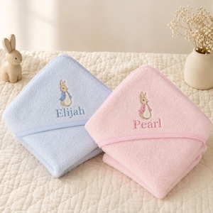 May include: Two hooded baby towels, one light blue and one light pink, each featuring an embroidered Peter Rabbit character and a personalised name. The blue towel is embroidered with the name Elijah, and the pink towel is embroidered with the name Pearl.