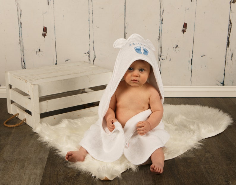 May include: A baby wrapped in a white hooded towel with blue embroidered footprints and the word "Baby". The baby is sitting on a white faux fur rug in front of a white wooden crate.