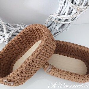 May include: Two oval-shaped storage baskets featuring a brown crocheted exterior with a thick, textured stitch pattern and a smooth, light-colored wooden base. Each basket includes a small tan leather-like tag with a silver metal rivet.