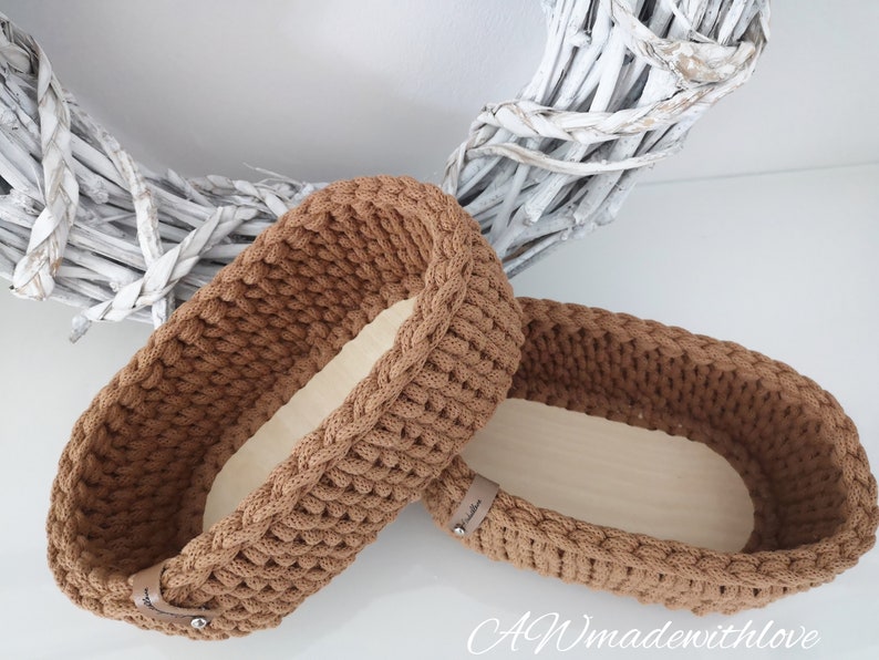 May include: Two oval-shaped storage baskets featuring a brown crocheted exterior with a thick, textured stitch pattern and a smooth, light-colored wooden base. Each basket includes a small tan leather-like tag with a silver metal rivet.