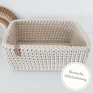 Puede incluir: Una cesta rectangular de ganchillo beige con un borde trenzado. La cesta tiene una base de madera de color claro y una pequeña etiqueta de cuero. La imagen también incluye una vista parcial de una decoración en forma de arcoíris en el fondo.