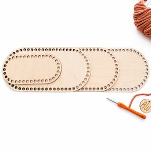 May include: Three light-colored oval wooden bases for crochet projects, each featuring a series of evenly spaced holes along the perimeter. The bases are stacked partially on top of one another, accompanied by a ball of orange yarn and a crochet hook.
