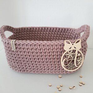 May include: A dusty rose oval crochet basket with integrated side handles. A light-colored wooden ornament shaped like a Christmas bauble with a bow and the letter A is attached to the front. A small fabric tag is sewn onto the upper left rim.