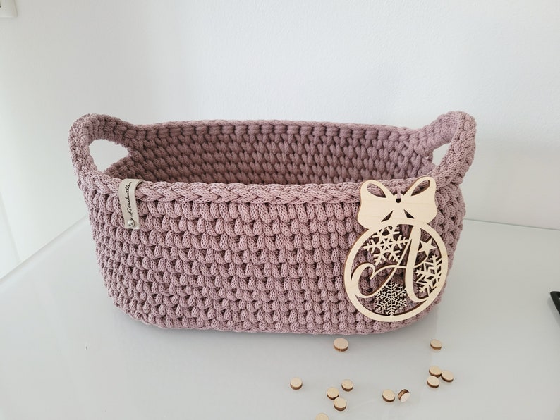 May include: A dusty rose oval crochet basket with integrated side handles. A light-colored wooden ornament shaped like a Christmas bauble with a bow and the letter A is attached to the front. A small fabric tag is sewn onto the upper left rim.