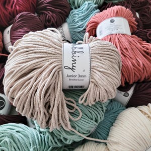 Könnte beinhalten: Eine Sammlung farbenfroher geflochtener Baumwollschnur-Spulen in verschiedenen Farbtönen, darunter Beige, Türkis und Burgunderrot. Das Etikett auf der vorderen Spule lautet "Junior 3mm Braided Cord". Die Schnüre sind eng gewickelt, was auf ihre Verwendung in Bastelprojekten hindeutet.