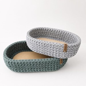 May include: Two oval-shaped storage baskets with thick, hand-crocheted walls in light gray and dark teal. Each basket features a smooth, light-colored wooden base and a small, rectangular brown leather tag attached to the side with a metal rivet.