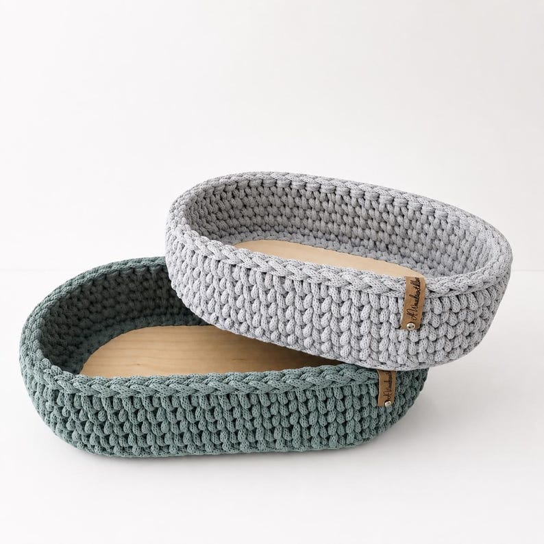May include: Two oval-shaped storage baskets with thick, hand-crocheted walls in light gray and dark teal. Each basket features a smooth, light-colored wooden base and a small, rectangular brown leather tag attached to the side with a metal rivet.