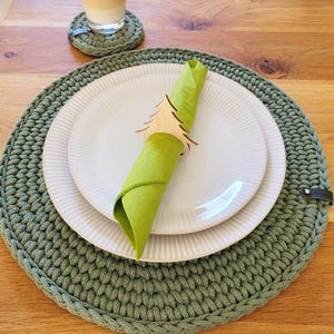 Op de afbeelding: Een groene gehaakte placemat met een witte plaat en een groene servet, vastgehouden door een houten boomvormige servetring. Een bijpassende onderzetter en een glas drank staan op de achtergrond.