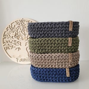 May include: A stack of four round, crocheted storage baskets in dark gray, olive green, beige, and navy blue. Each basket features a thick, textured knit pattern and a small, light brown leather-like tag attached with a silver metal rivet.