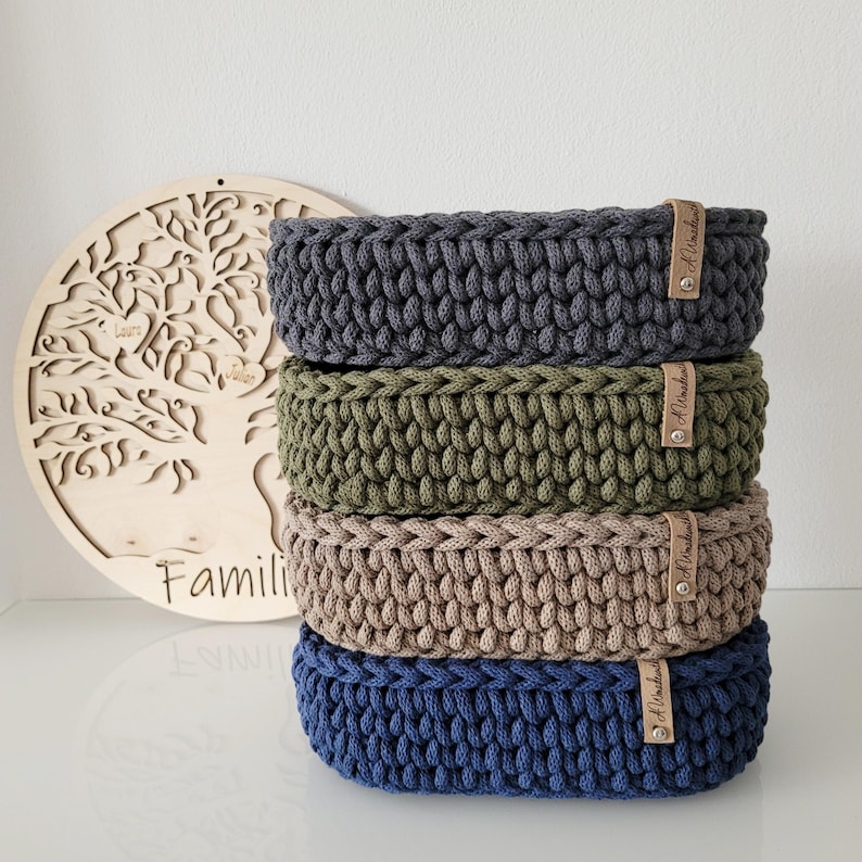 May include: A stack of four round, crocheted storage baskets in dark gray, olive green, beige, and navy blue. Each basket features a thick, textured knit pattern and a small, light brown leather-like tag attached with a silver metal rivet.