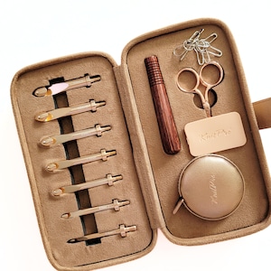 Può includere: Un kit da lavoro a maglia beige con vari strumenti. Il kit include uncinetti dorati, un ago di legno, piccole forbici dorate, graffette, un metro a nastro e un'etichetta in pelle con "KnitPro".