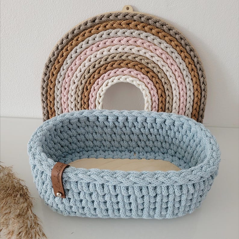 May include: A light blue oval crochet basket with a wooden base and a small brown leather tag featuring a metal rivet. Behind the basket sits a semi-circular wall hanging made of braided yarn in shades of beige, tan, pink, and cream.
