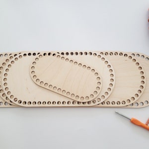 Könnte beinhalten: Vier ovale Häkelringe aus Holz in verschiedenen Größen, eine Häkelnadel und ein Holzschild mit den Buchstaben "AW" auf einer weißen Oberfläche.