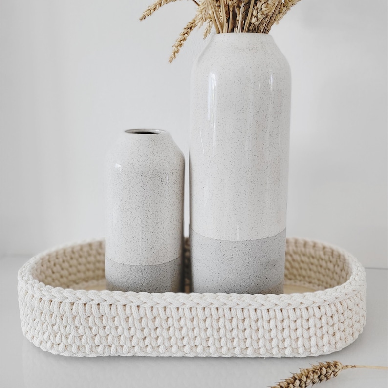 May include: Two cylindrical ceramic vases with a speckled white and gray finish, placed inside an oval-shaped, cream-colored crocheted basket. The taller vase contains a bundle of dried wheat stalks, adding a natural texture to the arrangement.