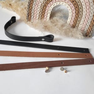 Könnte beinhalten: Ein schwarzer Ledergriff mit silbernen Nieten und drei Lederriemen in Schwarz, Hellbraun und Braun. Zwei silber- und goldfarbene Nieten sind ebenfalls sichtbar. Ein gewebter Regenbogen-Wandbehang in neutralen Tönen und getrocknetes Pampasgras befinden sich im Hintergrund.