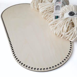Op de afbeelding: Een lichtgekleurde ovale houten mandbodem met voorgeboorde gaten langs de rand. Verschillende strengen off-white katoenen garen en een haaknaald liggen op de bodem, wat wijst op een knutselproject. De bodem is ongeveer 30 cm lang.