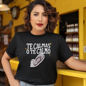 May include: Black t-shirt featuring the text "TE CALMAS O TE CALMO" and a flip-flop graphic. The person wearing the shirt has dark hair and gold hoop earrings.