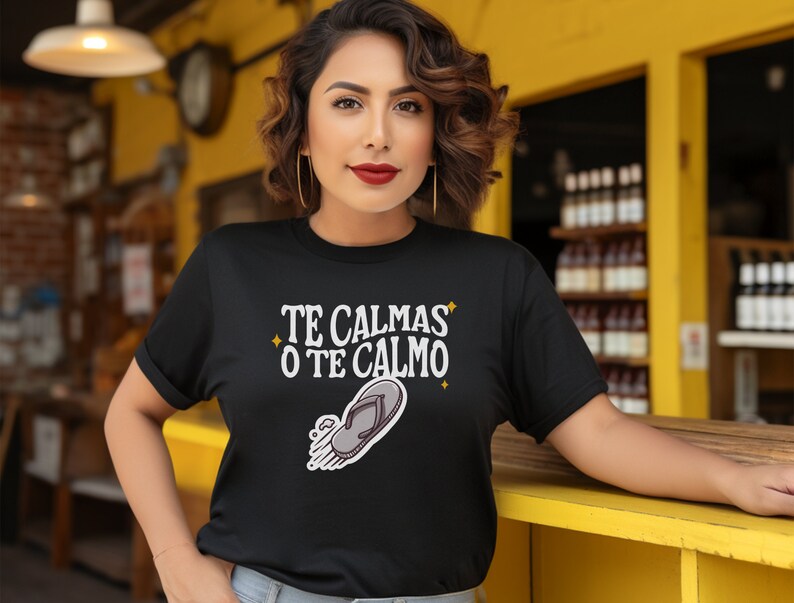 May include: Black t-shirt featuring the text "TE CALMAS O TE CALMO" and a flip-flop graphic. The person wearing the shirt has dark hair and gold hoop earrings.