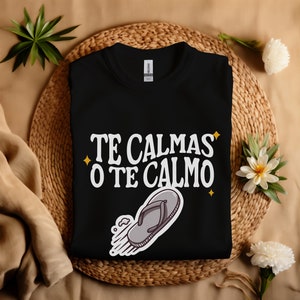 May include: Black crew neck t-shirt with the text "TE CALMAS O TE CALMO" in white, with a cartoon flip-flop graphic. The shirt is displayed on a woven mat with decorative greenery.