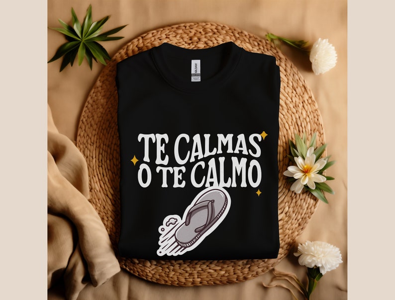 May include: Black crew neck t-shirt with the text "TE CALMAS O TE CALMO" in white, with a cartoon flip-flop graphic. The shirt is displayed on a woven mat with decorative greenery.