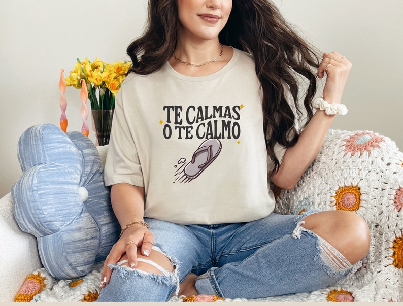 May include: Beige t-shirt with the text "TE CALMAS O TE CALMO" and a flip-flop graphic. The shirt is worn with ripped blue jeans. The image also shows a blue cushion, a vase of yellow flowers, and a white and pink crocheted blanket.