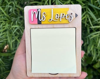May include: Wooden notepad holder with a pencil-shaped top that reads "Ms Lopez" in white script. The notepad is light-colored wood with a yellow, pink, and black pencil design. A pad of yellow sticky notes is in the center.