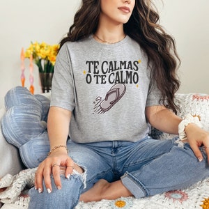 May include: A light grey t-shirt featuring the text "TE CALMAS O TE CALMO" and a flip-flop graphic. The shirt is worn by a person sitting on a chair. The person is wearing blue jeans.