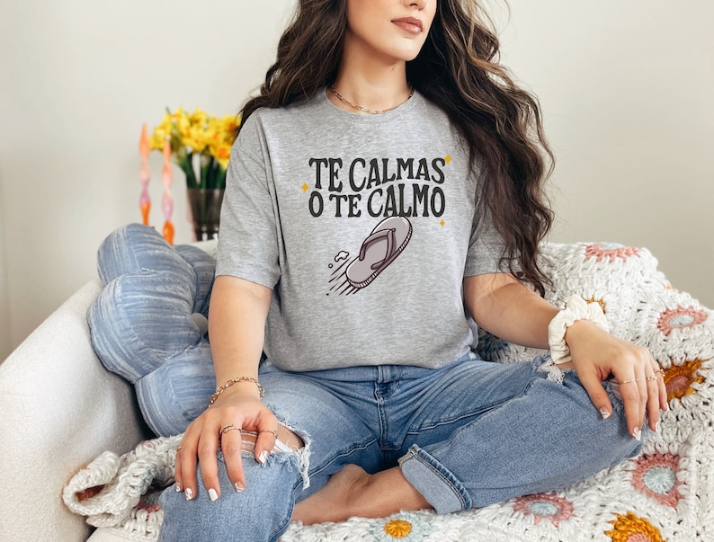 May include: A light grey t-shirt featuring the text "TE CALMAS O TE CALMO" and a flip-flop graphic. The shirt is worn by a person sitting on a chair. The person is wearing blue jeans.