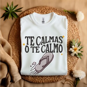 May include: White sweatshirt featuring the text "TE CALMAS O TE CALMO" in black and a flip-flop graphic. The garment is folded on a woven mat, decorated with flowers and greenery.