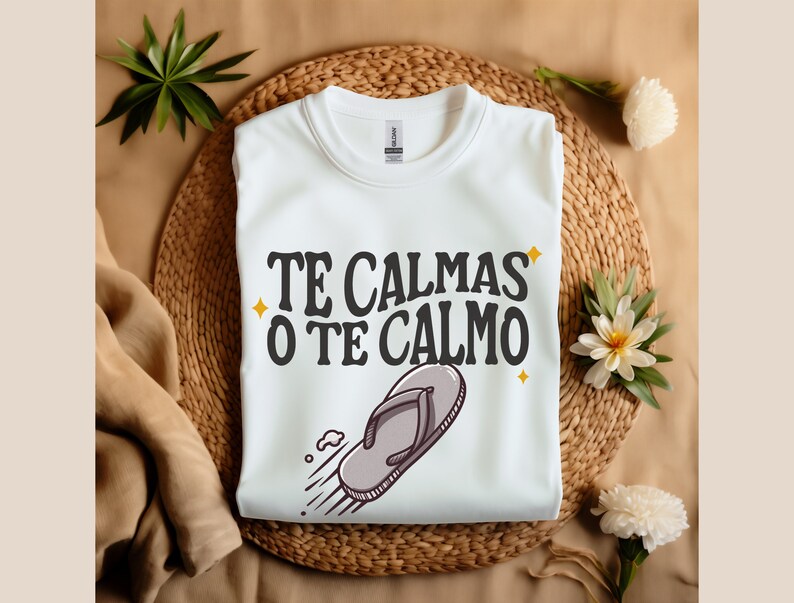 May include: White sweatshirt featuring the text "TE CALMAS O TE CALMO" in black and a flip-flop graphic. The garment is folded on a woven mat, decorated with flowers and greenery.