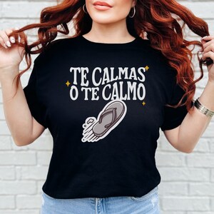 May include: Black t-shirt featuring the text "TE CALMAS O TE CALMO" in white, with a flip-flop graphic. The shirt has a crew neck and short sleeves. The text is in a stylized font with star accents.