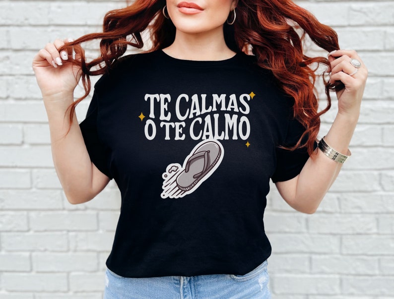 May include: Black t-shirt featuring the text "TE CALMAS O TE CALMO" in white, with a flip-flop graphic. The shirt has a crew neck and short sleeves. The text is in a stylized font with star accents.