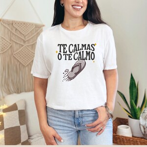 May include: White t-shirt featuring the text "TE CALMAS O TE CALMO" in black and a flip-flop graphic. The casual t-shirt is suitable for everyday wear.