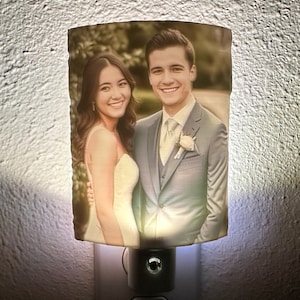 May include: A night light featuring a printed photograph of a smiling couple. The image is in sepia tones, with the couple dressed in formal attire. The night light is plugged into a wall outlet and emits a soft glow.