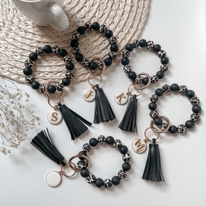May include: Five black and white patterned beaded bracelets with gold keychains and tassels. Each keychain has a white charm with a gold letter.
